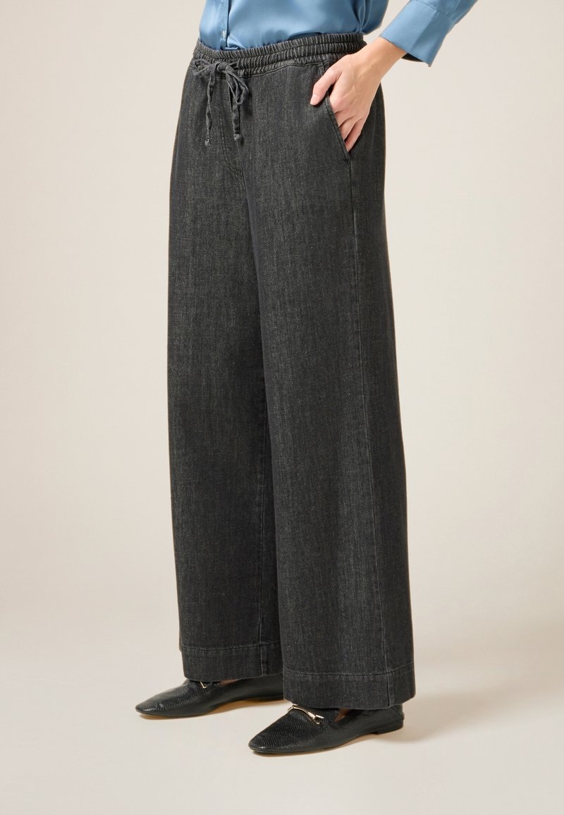 Weite schwarze Denim-Hosen mit elastischem Bund, Kordelzugdetails und Seitentaschen; kombiniert mit flachen schwarzen Schuhen.