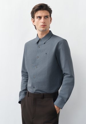 Mlad moški z rokami v žepih, oblečen v modro srajco z vzorcem in temno rjave hlače, rahlo gleda vstran.