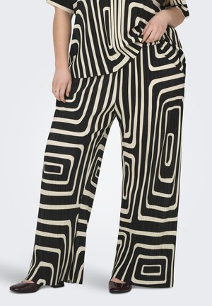 Pantalon à jambes larges noir et blanc avec motif géométrique audacieux, porté avec un haut assorti et des chaussures foncées.