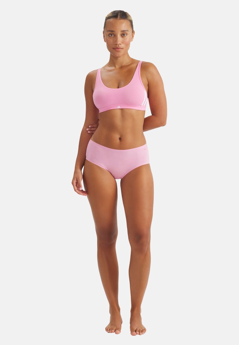 Mujer de pie descalza vistiendo un sujetador deportivo rosa y braguitas a juego, contra un fondo blanco liso.