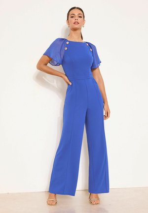 Femme portant une combinaison bleu cobalt avec des manches courtes transparentes et des boutons dorés, debout avec une main sur la hanche contre un fond blanc.