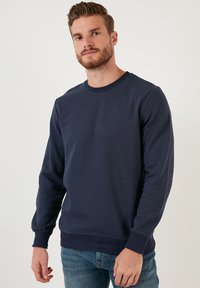 Námornícky modrý sveter s ribovanou textúrou, okrúhlym výstrihom a dlhými rukávmi. Obsahuje elastické manžety a lem pre tesné priliehanie.
