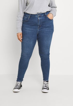 Person iført blå slim jeans, grå sweatshirt og sorte højtstående sneakers, stående med den ene hånd i lommen mod hvid baggrund.