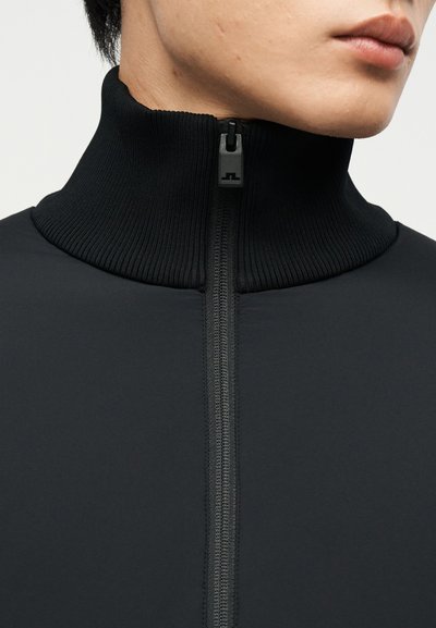 Κοντινή λήψη ενός ατόμου που φοράει μαύρο μπουφάν με φερμουάρ, με ραβδωτό ψηλό γιακά και εμφανές μαύρο φερμουάρ στο μπροστινό μέρος.