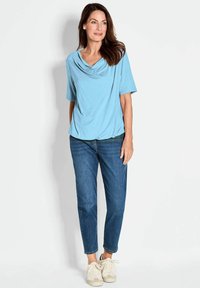 Lichtblauw kortmouwtop met een gedrapeerde halslijn, gecombineerd met medium-was jeans en off-white sneakers. Gladde, zachte stofstructuur.