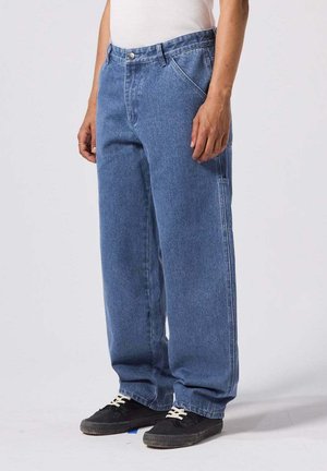 Persoon draagt losse blauwe spijkerbroek, wit tanktop en zwarte sneakers met witte veters, staand tegen een effen achtergrond.