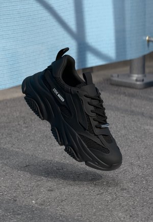 Black Steve Madden sneaker with mesh and leather details, thick sole, floating above textured asphalt next to light blue wall with shadows.