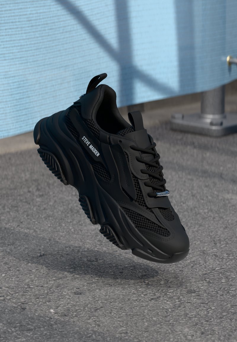 Baskets noires Steve Madden avec des détails en mesh et cuir, semelle épaisse, flottant au-dessus d'un asphalte texturé à côté d'un mur bleu clair avec des ombres.