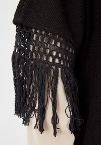 Tejido de punto negro con intrincados detalles de flecos a lo largo de la manga, que revela una sutil capa beige por debajo de los acentos de ganchillo.
