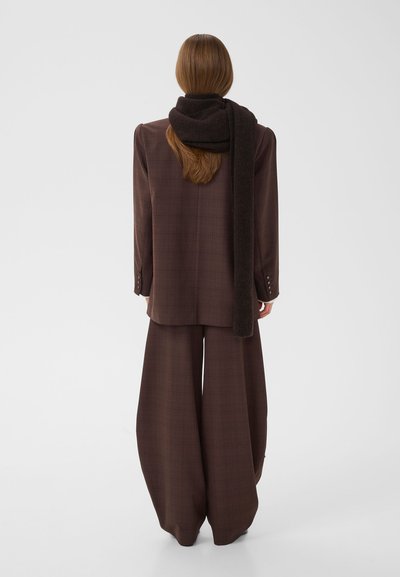 Femme aux longs cheveux porte un costume à carreaux marron oversize et une écharpe marron foncé, debout dos à un fond clair uni.