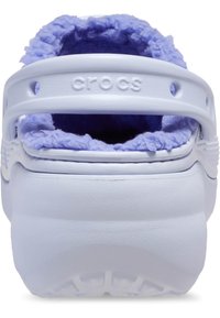 Des Crocs blancs avec une doublure en laine de couleur lavande. L'arrière dispose d'une sangle et d'un logo en relief. La semelle texturée indique une bonne adhérence.