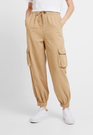 Pantalon cargo beige avec taille élastique, cordon de serrage, poches latérales à rabat et poignets élastiques, porté avec des baskets blanches et un top court blanc.