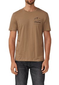 Mann trägt ein braunes T-Shirt mit kurzen Ärmeln, schwarzem Bergmotiv und Text auf der linken Brust, kombiniert mit dunklen Jeans.