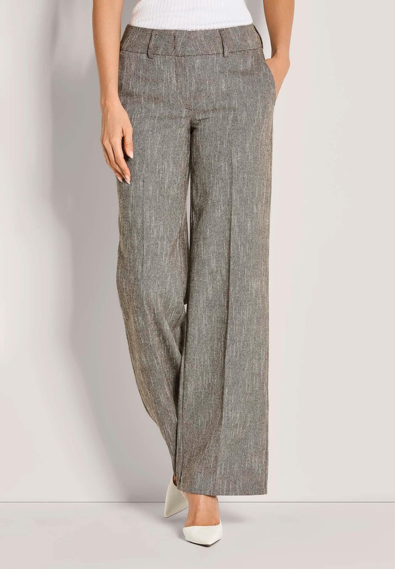 Vrouw in hoge taille, wijde grijze textuurbroek, gecombineerd met een witte ribbelige mouwloze top en witte puntige hakken.