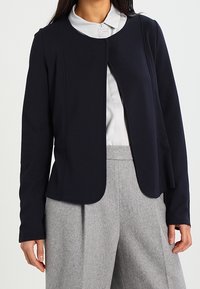 Laivastonsininen takki, joka on valmistettu sileästä kankaasta, pyöreällä pääntiellä, edestä suljettava ja räätälöidyllä istuvuudella, yhdistetty vaaleanharmaisiin housuihin.