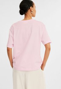 Ljust rosa kortärmad t-shirt med avslappnad passform, rund halsringning och mjuk bomullstextur, bärs med beige byxor.