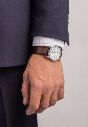 Montre en métal argenté avec un cadran blanc, des chiffres noirs et un bracelet en cuir marron, portée sur une main dans un costume bleu marine ajusté.