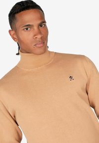 Beige coltrui gemaakt van zacht materiaal. Heeft een geribde kraag en manchetten. Klein geborduurd logo op de linkerkant van de borst. Glad oppervlak.
