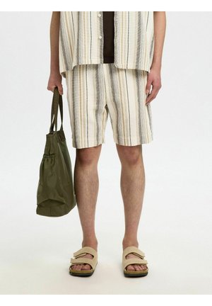 Osoba oblečená v béžových pruhovaných kraťasoch a košeli, nosí béžové sandále, vedľa seba drží olivovo zelenú tašku na rameno.