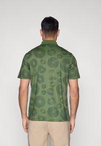 Žalia polo marškinėliai su tekstūruotu gėlių raštu tamsesnių žalių atspalvių, trumpomis rankovėmis ir klasikiniu apykakliu. Paprastas nugaros dizainas.