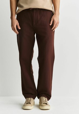 Man wearing brown relaxed-fit trousers and beige sneakers, standing against a plain light background.