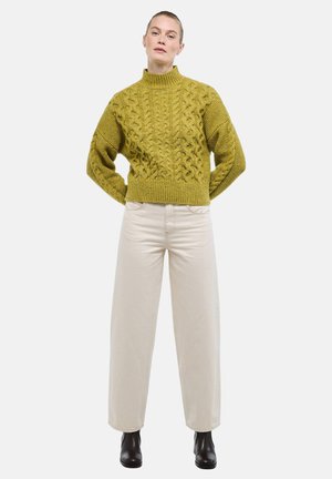 Femme portant un pull en maille torsadée moutarde, un pantalon large écru et des bottines noires, debout les mains dans le dos.