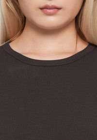 Nærbilde av en kvinnes nedre ansikt, nakke og øvre bryst iført en mørk ribbet skjorte med glansfulle lepper synlige.