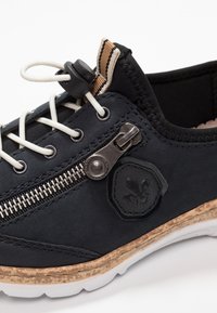 Baskets noires avec une tige lisse, des lacets blancs, une fermeture éclair sur le côté et une semelle en caoutchouc texturée. Présente un écusson de logo et des accents contrastants.