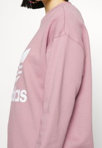 Persona vistiendo una sudadera Adidas de color púrpura claro, vista de perfil, con cabello corto negro y aretes coloridos visibles.