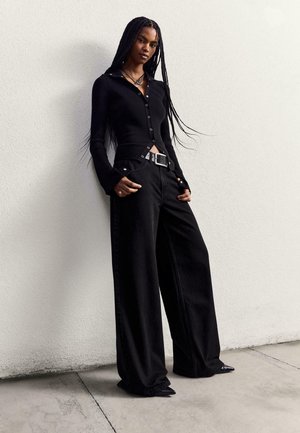 Schwarzes geripptes Langarmoberteil mit Knöpfen und hochgeschnittenen schwarzen weit geschnittenen Hosen, die mit einem silbernen Gürtel mit rechteckiger Schnalle versehen sind.