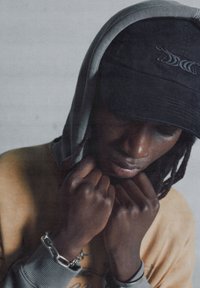 Szürke kapucnis pulóver és fekete sapka hímzett mintával, közelkép a kezektől, amelyek az arc közelében összefonódnak. Puha szövet textúra, sötét bőrszín, finom lánc részlet.