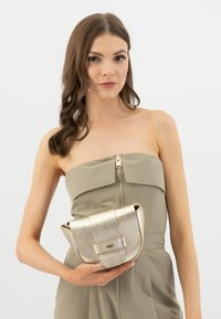 Gouden metallic handtas met een textuurafwerking, gebogen vorm, klep sluiting en een voorste sluiting, gehouden tegen een lichte achtergrond.