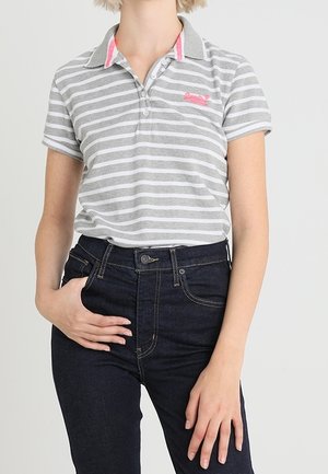Femme portant un polo gris et blanc à rayures avec des détails de col roses et un jean taille haute foncé, debout avec une main dans la poche.