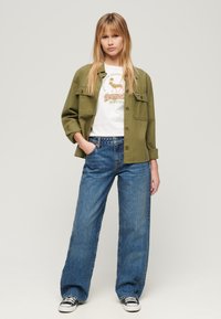 Oliwkowa kurtka zapinana na guziki nałożona na białą koszulkę z grafiką, w zestawieniu z szerokimi niebieskimi dżinsami i czarno-białymi sneakersami.