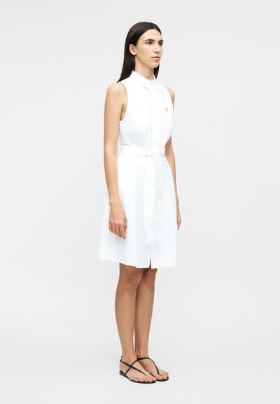 Nő áll egy ujjatlan, fehér, gombos ruhában, övvel, fekete lapos szandállal, egy egyszerű szürke háttér előtt.