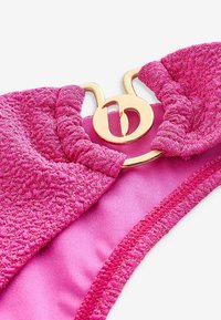 Růžový bikini top s texturovanou tkaninou a metalickým vláknem, zdobený zlatým kruhovým kovovým detailem uprostřed.