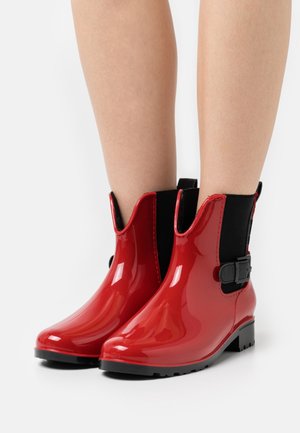 Glinsterende rode enkellaarsjes met zwarte elastische zijkanten en gespsluiting, gedragen op blote benen, geplaatst tegen een witte achtergrond.