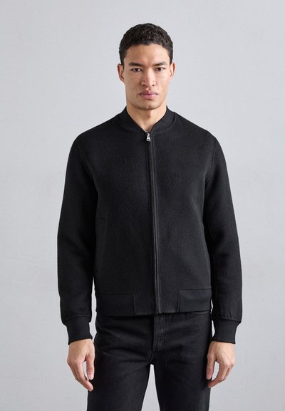 Veste bomber noire zippée, design ajusté, poignets et ourlet côtelés, en tissu texturé, avec poches latérales, portée sur un jean sombre.