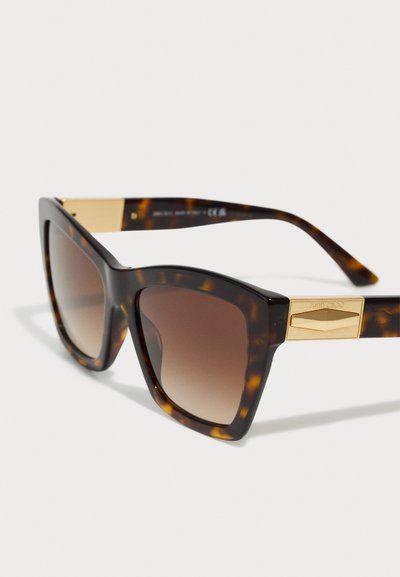 Lunettes de soleil en écaille de tortue avec des verres dégradés marron, des montures carrées audacieuses, et des détails dorés sur les branches. Matériau plastique durable.