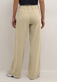 Ljusbeige vida byxor med elastisk midja, tillverkade av ett mjukt, strukturerat tyg. Fållarna är prydliga med en lätt avsmalning i botten.