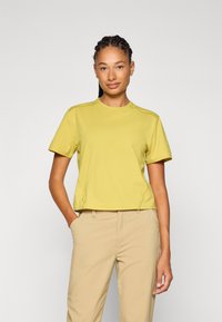 T-shirt giallo a maniche corte con scollo rotondo, caratterizzato da dettagli sulle cuciture laterali e una vestibilità rilassata, abbinato a pantaloni beige chiaro.