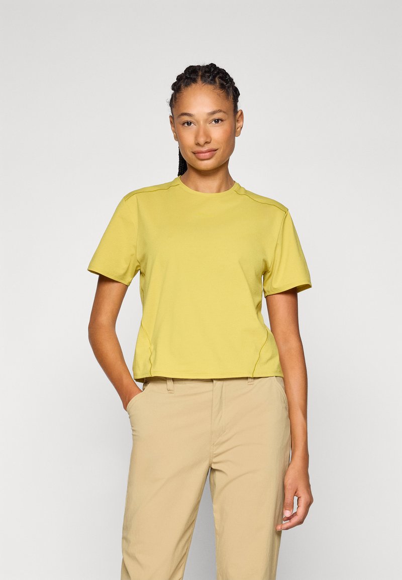 T-shirt giallo a maniche corte con scollo rotondo, caratterizzato da dettagli sulle cuciture laterali e una vestibilità rilassata, abbinato a pantaloni beige chiaro.