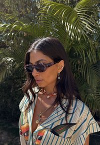 Firkantede solbriller i et pink skildpaddemønster. Personen har en stribet skjorte med farverige fugleprint og lagdelte halskæder på.