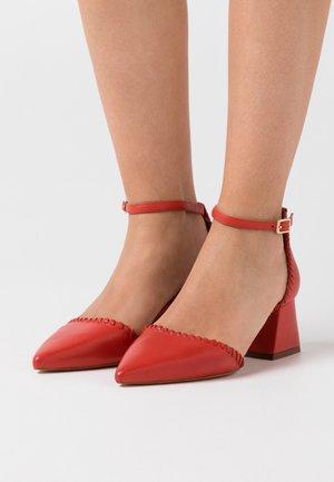 Pieds portant des escarpins rouges à talons blocs pointus avec des brides autour de la cheville et des coutures décoratives le long des bords sur fond blanc.