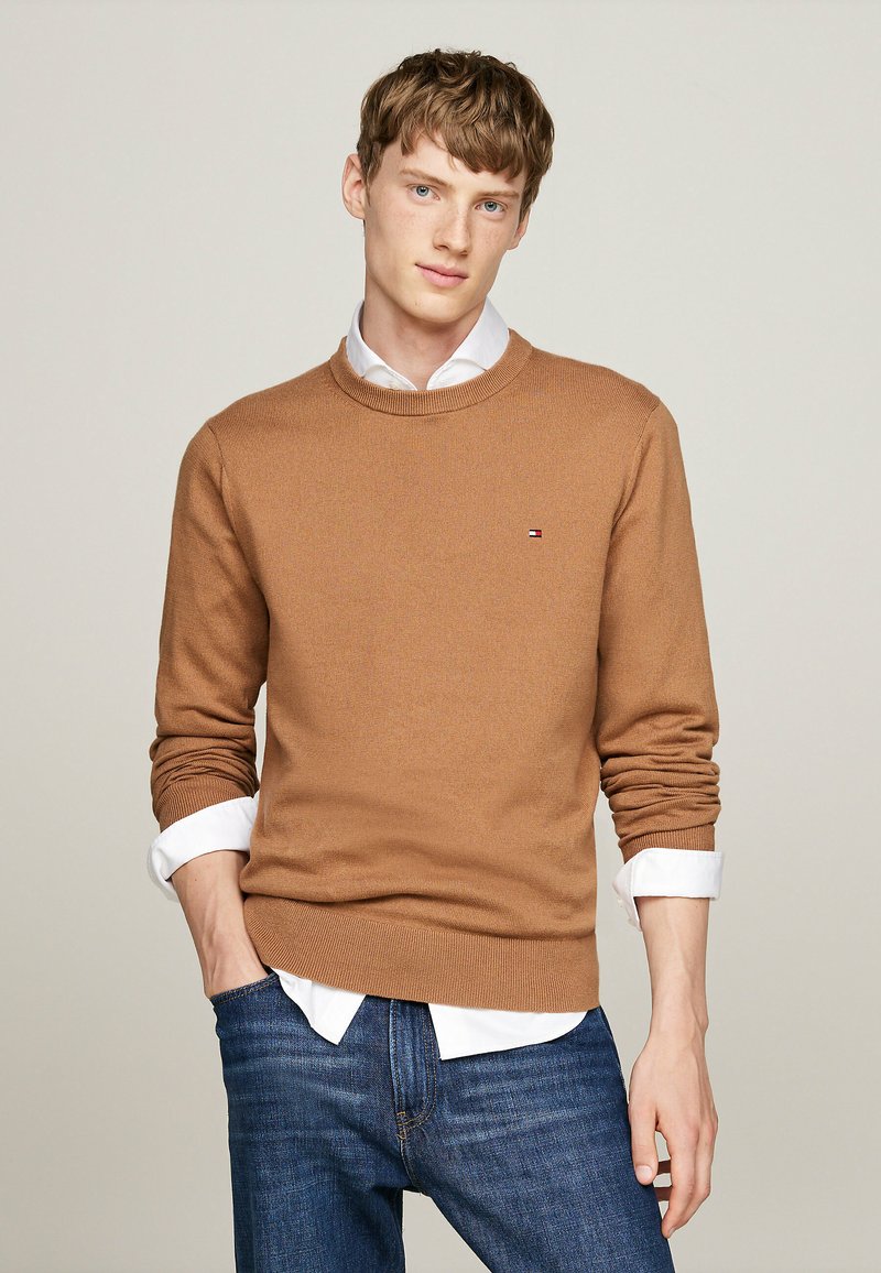 Brauner Strickpullover mit rundem Ausschnitt, kombiniert mit einem weißen Hemd mit Kragen. Die Person trägt eine blaue Jeans.