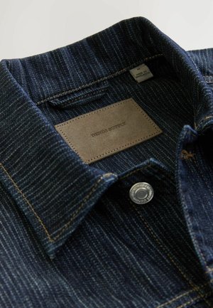 Denim overhemd met een donkerblauwe, gestructureerde stof, een kraag, een leren label met de tekst "DENIM SUPPLY" en een zilveren drukknoop.