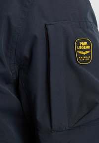 Tela de chaqueta azul oscuro con un parche prominente amarillo y negro que presenta "PME LEGEND" y "AMERICAN CLASSIC", ubicado en la parte superior de la manga.