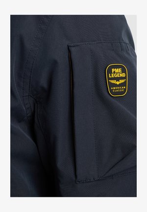 Tela de chaqueta azul oscuro con un parche prominente amarillo y negro que presenta "PME LEGEND" y "AMERICAN CLASSIC", ubicado en la parte superior de la manga.
