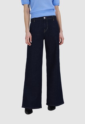 Femme portant un jean bleu foncé à jambes larges et un pull léger bleu clair à manches courtes, debout devant un fond uni.