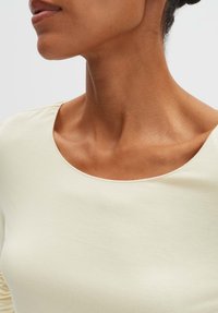 Lichtbeige top gemaakt van zachte stof, met een brede, ronde halslijn en een gladde textuur, met zichtbare huidskleur eronder.